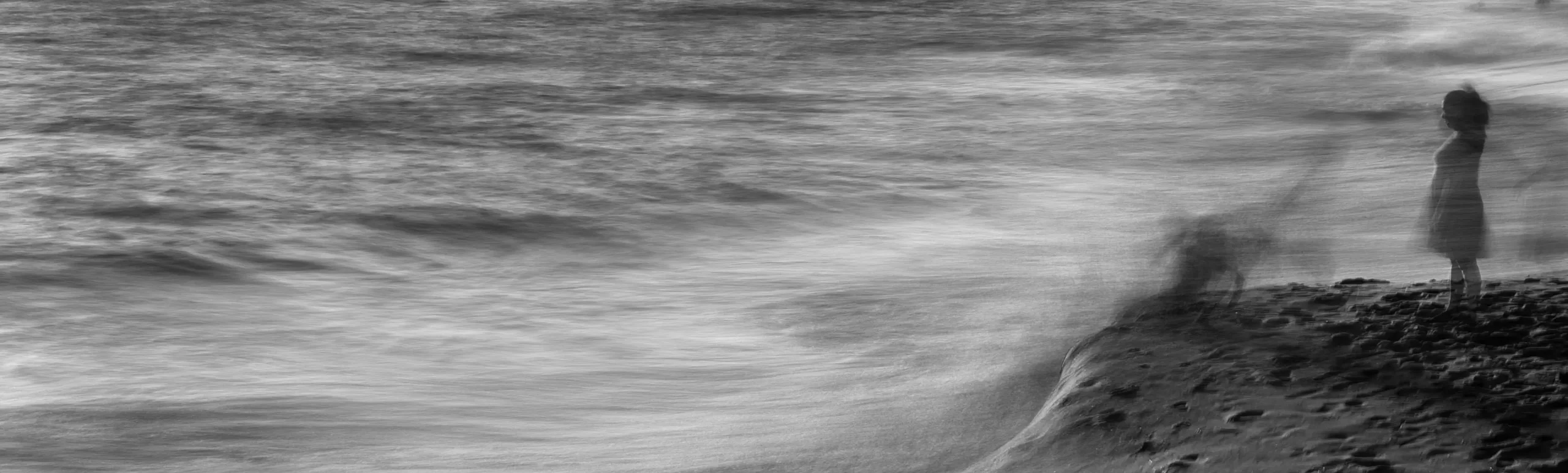 Long exposure black and white photograph of a ghostly blurred figure standing on a sandy beach beside a turbulent sea.