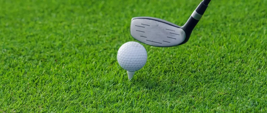 A golf club head positioned above a white golf ball on a tee, ready to swing on a manicured fairway.