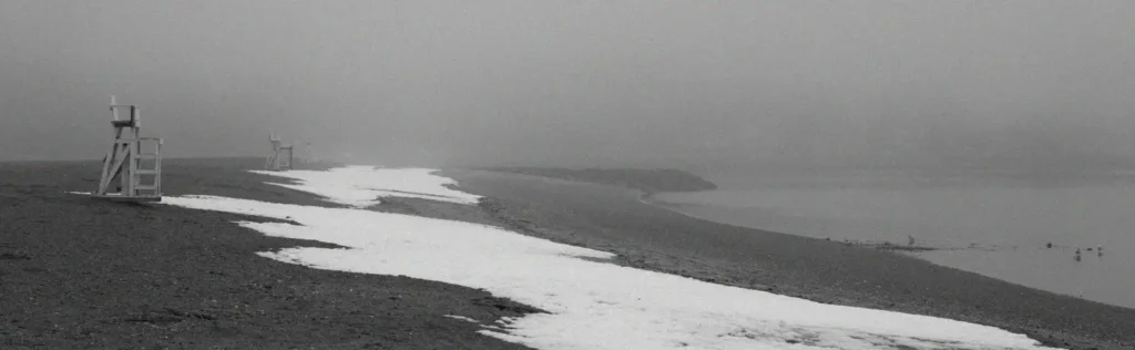 Foggy, desolate beach scene in black and white with snow or heavy mist covering parts of the sand and lifeguard towers visible in the distance.
