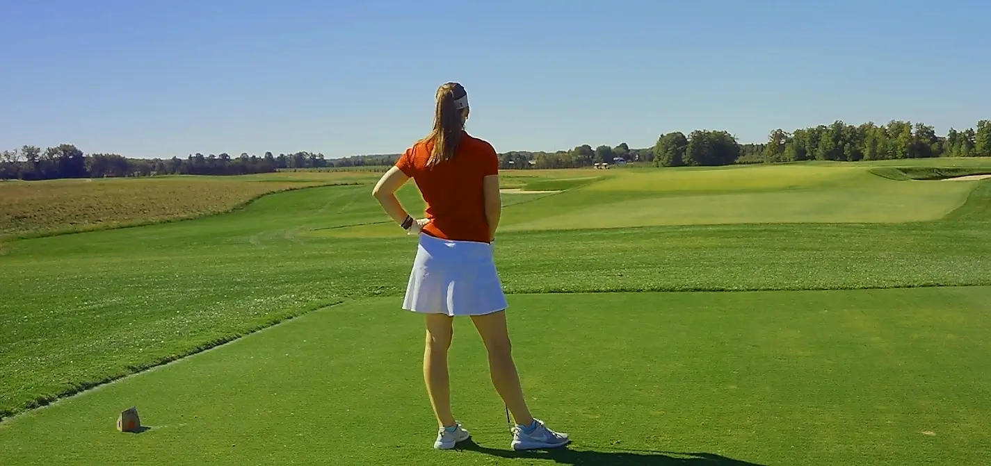 A woman in an orange shirt and white skirt stands on a golf tee box with her hands on her hips, looking out over a beautiful expansive golf course on a sunny day.