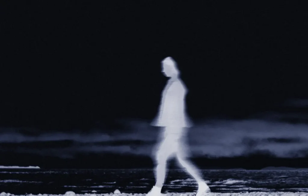 Motion-blurred ghostly figure walking along a dark beach at night, creating an ethereal and mysterious atmosphere