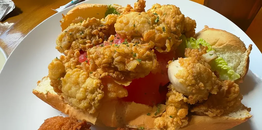 Close-up of a loaded fried shrimp sandwich on a soft bun, overflowing with golden crispy fried shrimp, fresh tomato slices, lettuce, and garnished with chopped parsley.
