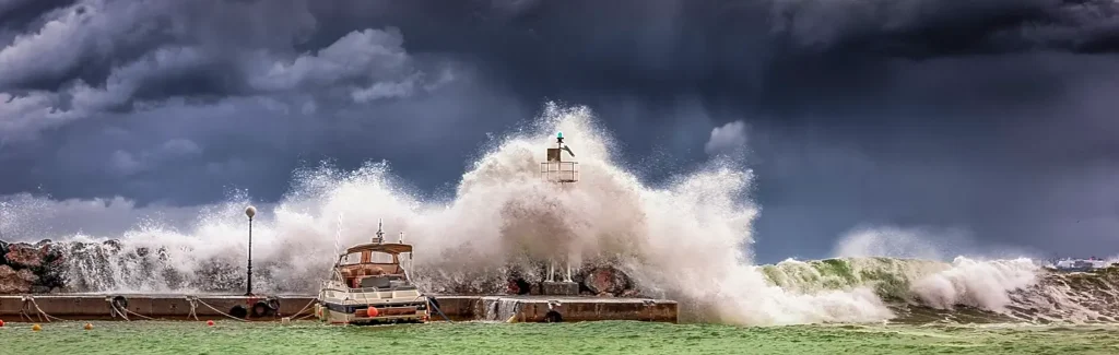 Dramatic scene of massive waves crashing violently over a harbor pier during a storm, sending huge sprays of water into the air around a boat under dark, ominous clouds.