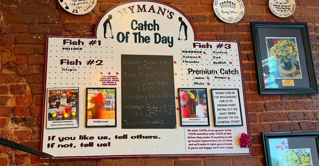 Wall-mounted menu board at Hyman's Catch Of The Day restaurant. It lists the Fish of the Day (Pollock, Tilapia, Haddock, etc.), specialty cocktails like Bloody Mary and Moonshine Margarita, and various promotions. The sign is mounted on a red brick wall with decorative plates and framed pictures.