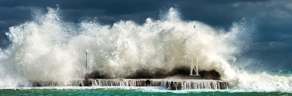 A giant wave exploding against a pier or breakwater, creating a massive wall of white spray that engulfs a small lighthouse-like structure during a fierce storm.