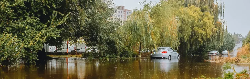 Flooded residential street after a storm, with a silver car partially submerged in deep brown floodwater among trees and buildings.