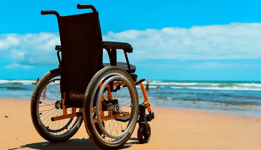 A black and orange wheelchair parked on a sunny sandy beach, facing the ocean under a bright blue sky with scattered clouds. People are visible swimming in the background.