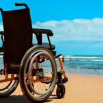 A black and orange wheelchair parked on a sunny sandy beach, facing the ocean under a bright blue sky with scattered clouds. People are visible swimming in the background.
