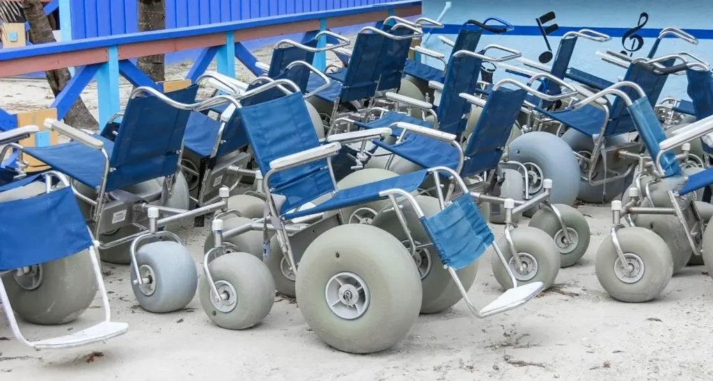 Multiple blue beach wheelchairs with large balloon tires lined up on the sand, ready for use near a blue beach structure.