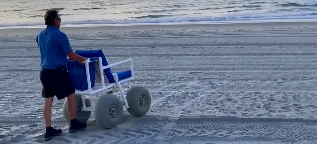 A man in a blue shirt pushing a blue beach wheelchair with large tires across the sand, leaving tracks behind him near the shoreline.