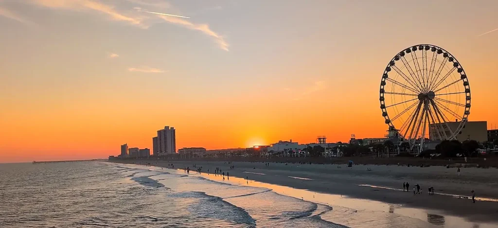 Stunning sunset over Myrtle Beach with the giant SkyWheel Ferris wheel on the right, a long sandy beach with people, ocean waves, and vibrant orange sky.