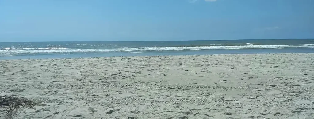 Expansive sandy beach with footprints scattered across the light-colored sand, leading to the ocean where gentle waves with white foam are breaking under a clear blue sky.