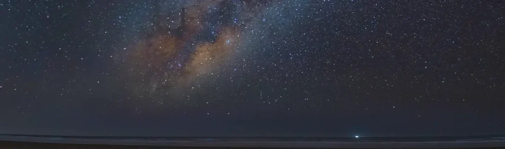 Milky Way visible above the ocean horizon with a faint light on the beach below