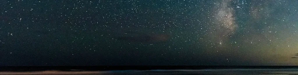 Milky Way glowing above a dark beach horizon with visible cloud cover