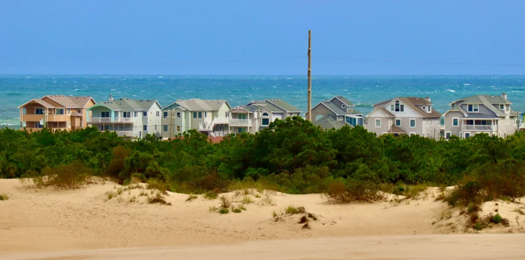 Row of colorful beach houses along the oceanfront in Nags Head, North Carolina, with sand dunes and sea oats in the foreground under a clear blue sky.