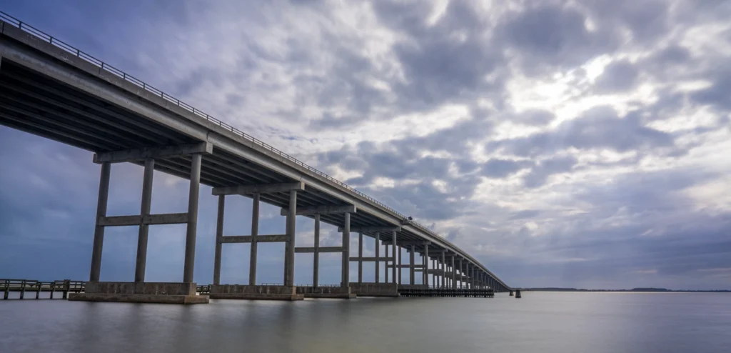 Long concrete highway bridge stretching across calm water under a dramatic cloudy sky, supported by numerous pillars