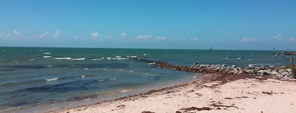 Sunny beach scene with light-colored sand in the foreground and a rocky jetty extending into the turquoise ocean. Gentle waves roll toward the shore, with a person swimming in the water and navigation markers visible in the distance under a bright blue sky.