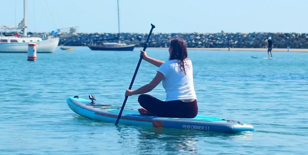 A woman in a white t-shirt sitting on a blue inflatable paddleboard labeled 'PERFORMER 11' while paddling on calm water, with sailboats and a rocky shoreline in the background.