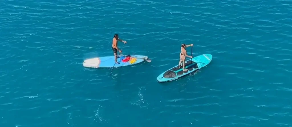Aerial view of a man and a woman each standing on separate paddleboards, paddling across bright turquoise water.