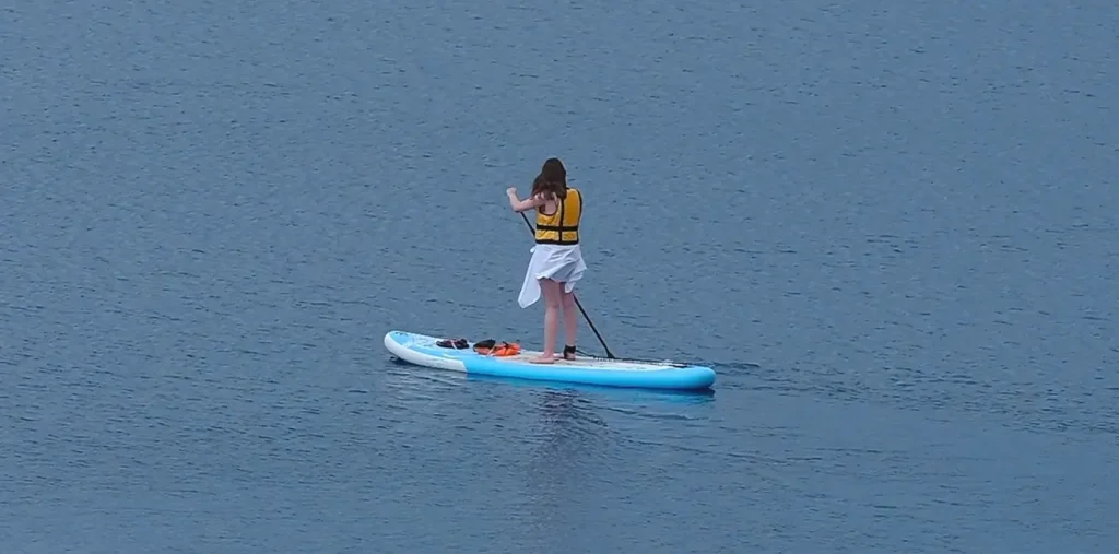 A woman wearing a yellow life vest and white skirt standing on a blue and white paddleboard while paddling on calm blue water, viewed from above.