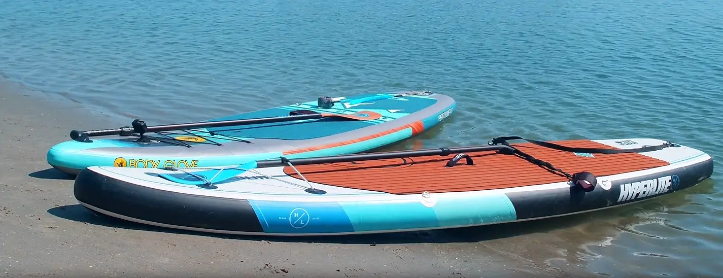 Two inflatable paddleboards resting on a sandy beach by the water: a black and turquoise Hyperlite board in the foreground and a turquoise and gray Body Glove board behind it.