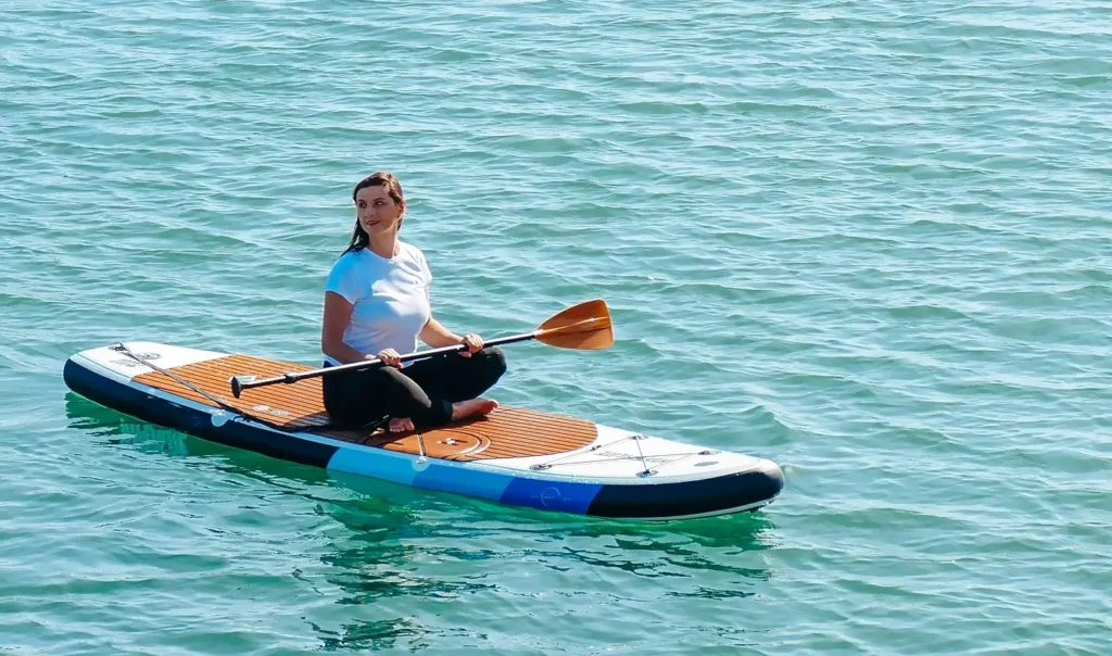 A woman sitting cross-legged on a paddleboard in calm turquoise water, holding a wooden paddle and smiling at the camera on a sunny day.