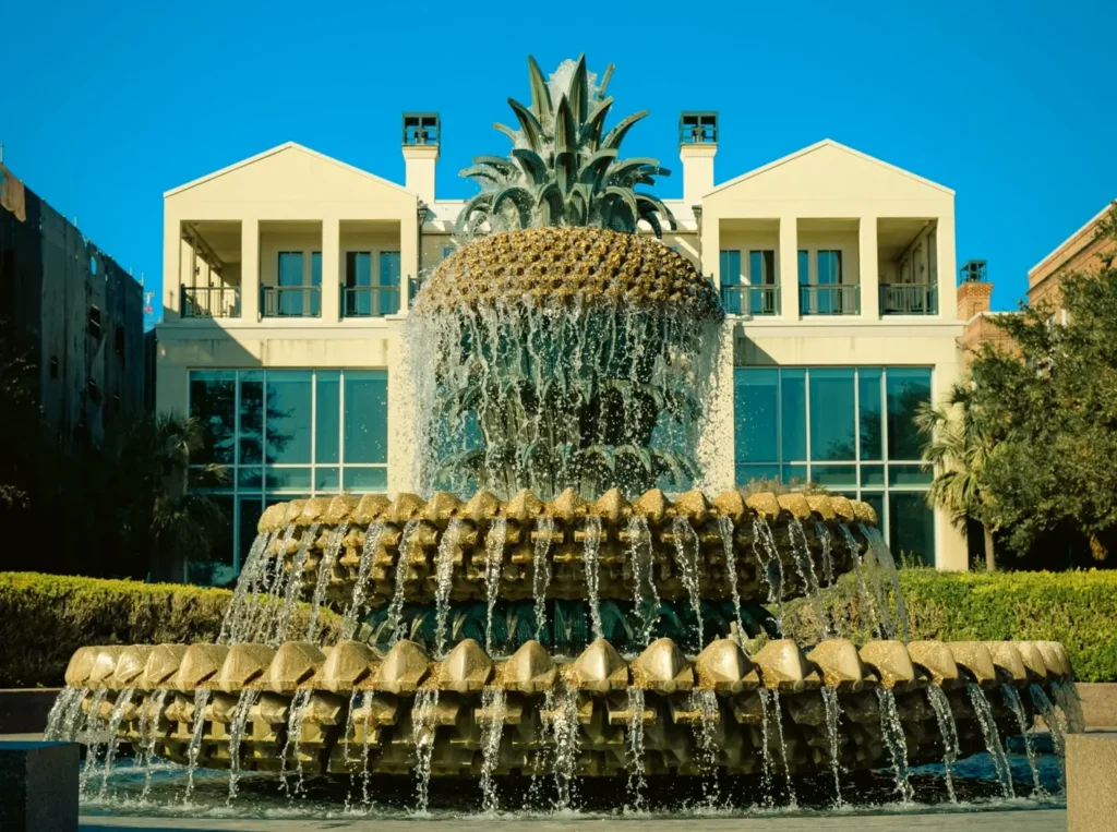 Iconic Pineapple Fountain in Waterfront Park, Charleston, with water cascading from the large bronze pineapple sculpture, set against white buildings on a sunny day.