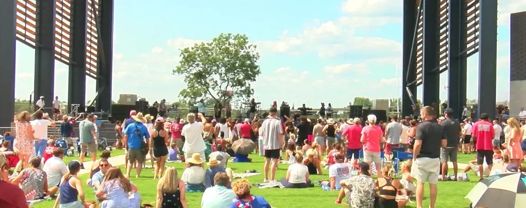 Large crowd enjoying a sunny outdoor concert on the grassy lawn at Live Oak Bank Pavilion at Riverfront Park, with the stage visible in the background.