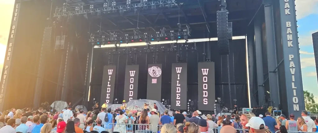 Large concert stage at Live Oak Bank Pavilion with "Wilder Woods" banners during a daytime performance, packed with audience members.