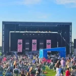 Wide shot of a full crowd at Live Oak Bank Pavilion during a sunny afternoon concert, showing the large stage and extensive lawn seating area.