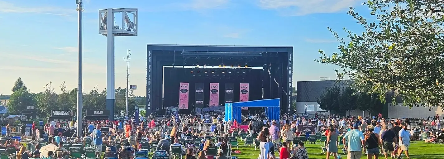 Wide shot of a full crowd at Live Oak Bank Pavilion during a sunny afternoon concert, showing the large stage and extensive lawn seating area.