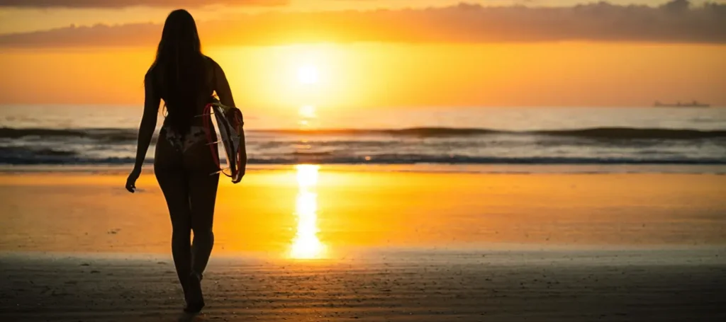 Silhouette of woman walking beach at sunset carrying surfboard