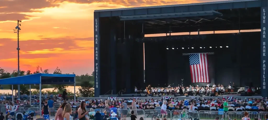 Beautiful sunset concert at Live Oak Bank Pavilion with a full orchestra performing on stage and an American flag displayed, as the audience watches from the lawn.