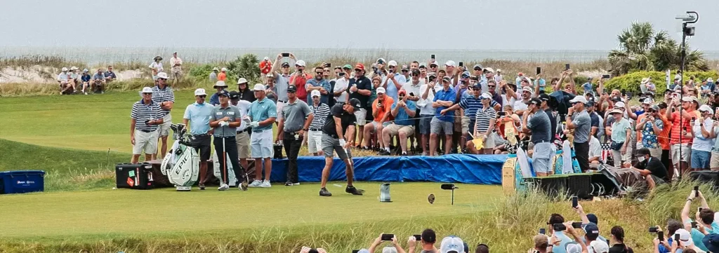 Spectators watching a golfer tee off near the ocean at a coastal golf tournament