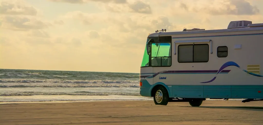 A large white and blue Class A RV parked on the beach at golden hour with ocean waves rolling in, showcasing oceanfront RV camping at its best.