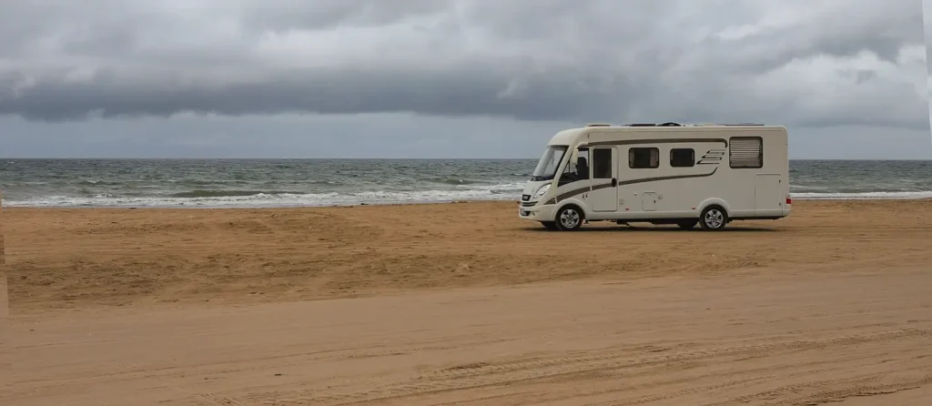 A white Class C motorhome parked on a wide sandy beach under dramatic cloudy skies, with the ocean in the background — perfect coastal RV scene.