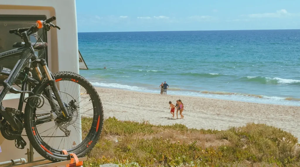 View from an RV campsite with a mountain bike mounted on the back, overlooking a sandy beach where a family plays near the ocean on a sunny day.