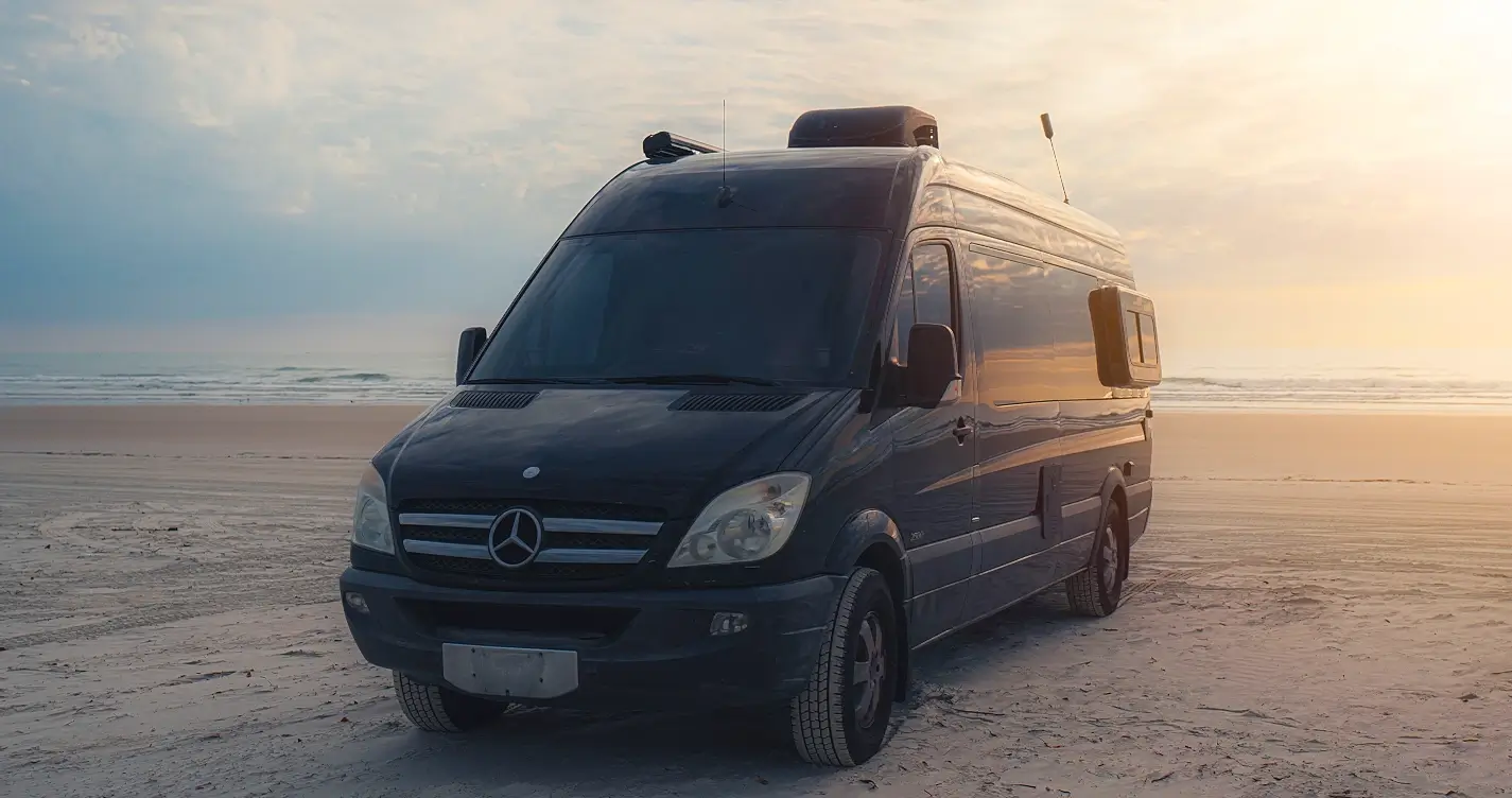 A sleek black Mercedes Sprinter camper van parked directly on the beach at sunset, with soft ocean waves in the background