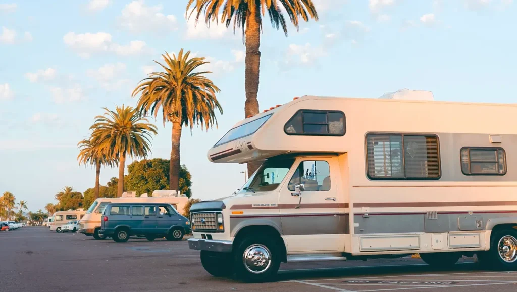 A vintage white and brown Class C RV parked in a sunny lot lined with tall palm trees under a blue sky, evoking classic coastal RV travel.