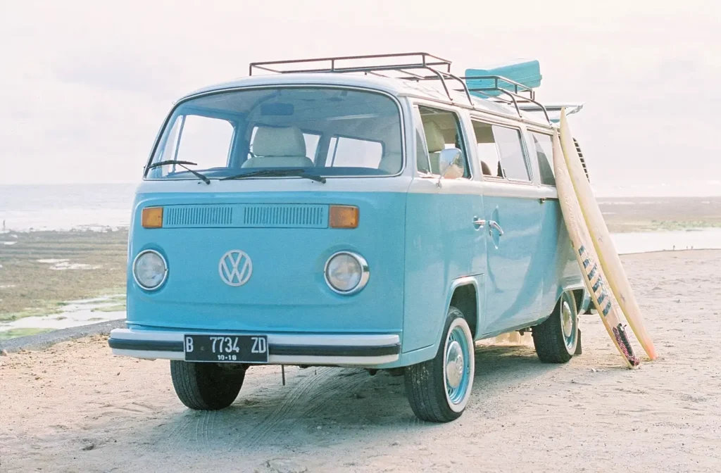 A classic light blue Volkswagen Type 2 camper van parked on the sand at the beach with surfboards leaning against it, capturing the spirit of beach van life.