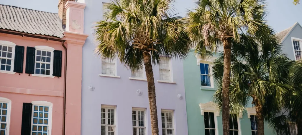 Colorful pastel historic buildings in shades of pink, purple, and teal with black shutters line a sunny street, framed by tall palm trees under a clear blue sky.