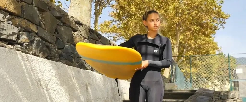 A young woman in a black wetsuit holds a bright yellow surfboard under her arm while standing outdoors near a stone wall on a sunny day.