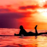Silhouette of a woman lying on a surfboard in the ocean during a vibrant red and orange sunset, with one leg raised