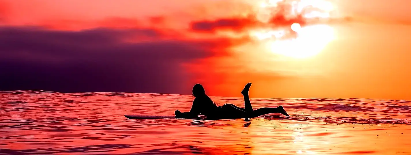 Silhouette of a woman lying on a surfboard in the ocean during a vibrant red and orange sunset, with one leg raised