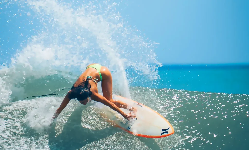 Female surfer in a bright green bikini crouched low on her surfboard, riding a powerful wave with a massive splash of white water behind her against a clear blue sky.