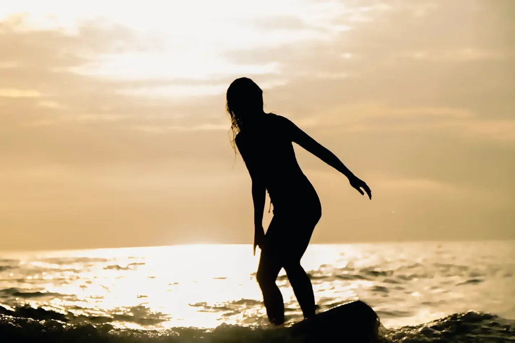 Silhouette of a woman surfing at sunset, standing on her board with the golden sun reflecting on the ocean water behind her.