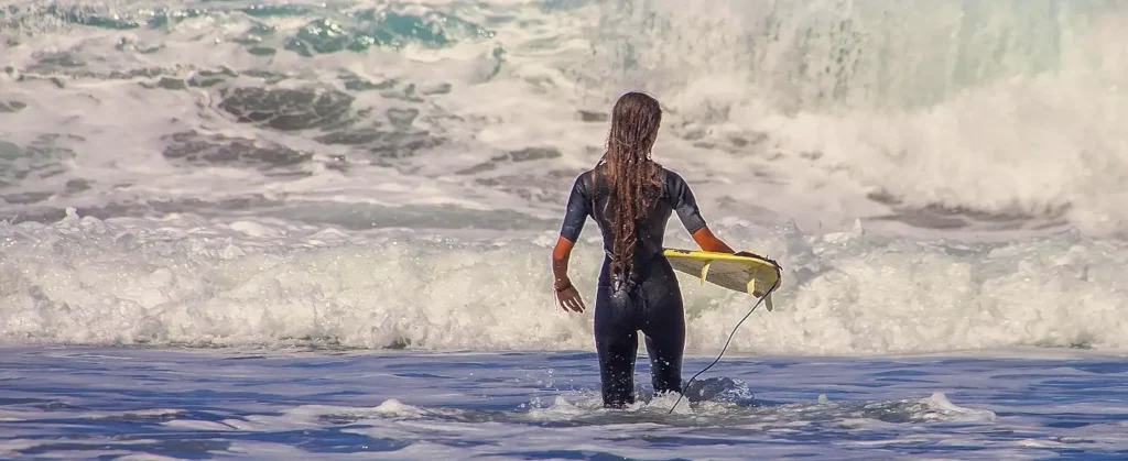 A female surfer in a black wetsuit stands in the water with her back to the camera, holding a yellow surfboard while facing large crashing waves