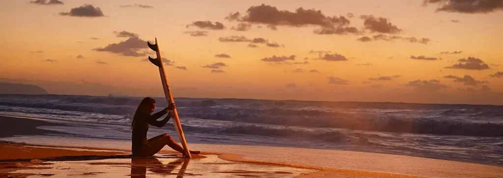 A woman in a full wetsuit sits on the wet sand at sunset, holding her surfboard upright beside her while gazing at the ocean waves.