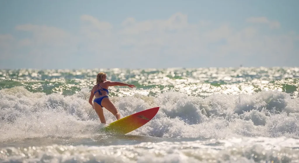 Woman in a blue bikini balancing on a red and yellow surfboard while riding a wave, with bright sunlight reflecting off the ocean surface.