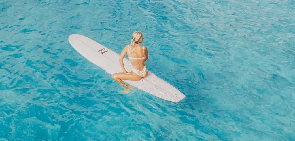Blonde woman in a white bikini sitting on a white surfboard in calm, clear turquoise water, viewed from above.
